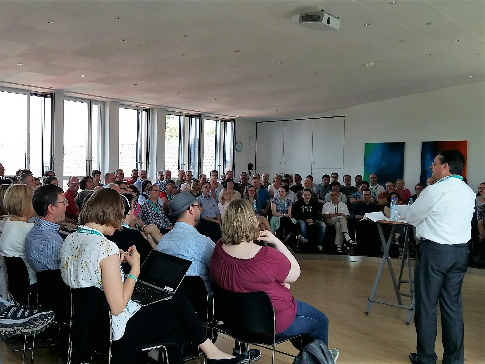 Eine Person steht vor einem Publikum in einem Raum mit vielen Sitzplätzen, während sie eine Präsentation hält.