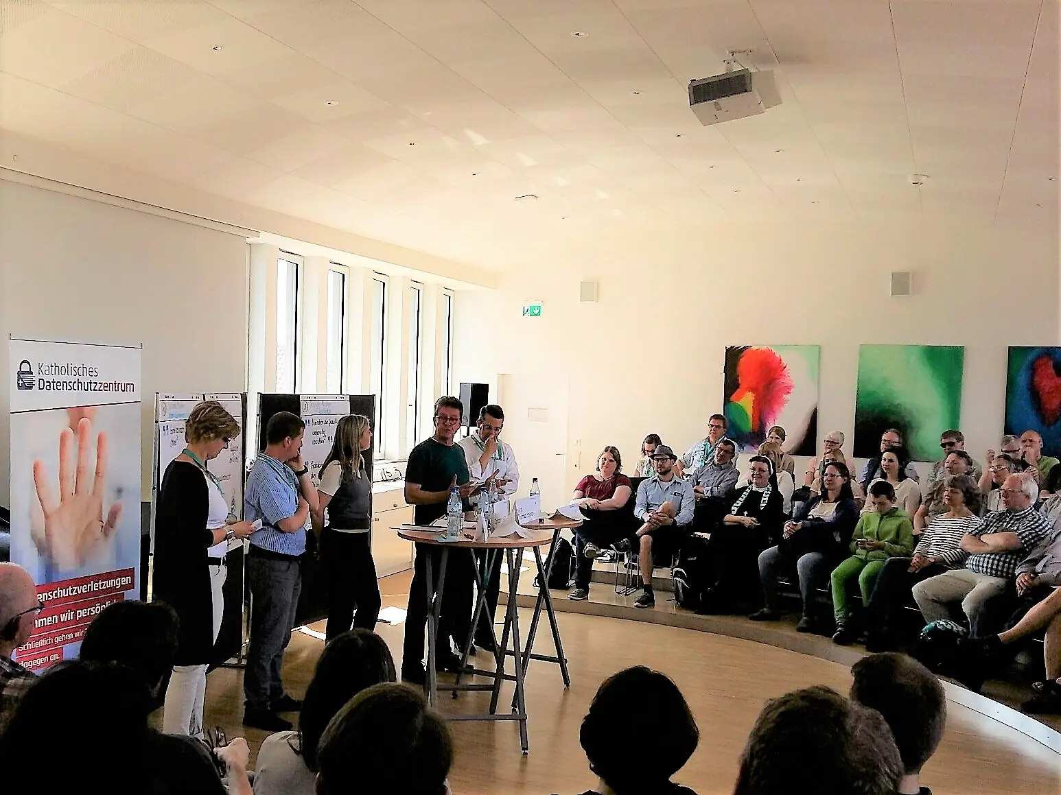 Gruppendiskussion mit mehreren Personen auf einer Bühne, während Publikum in einem hellen Raum zuschaut.