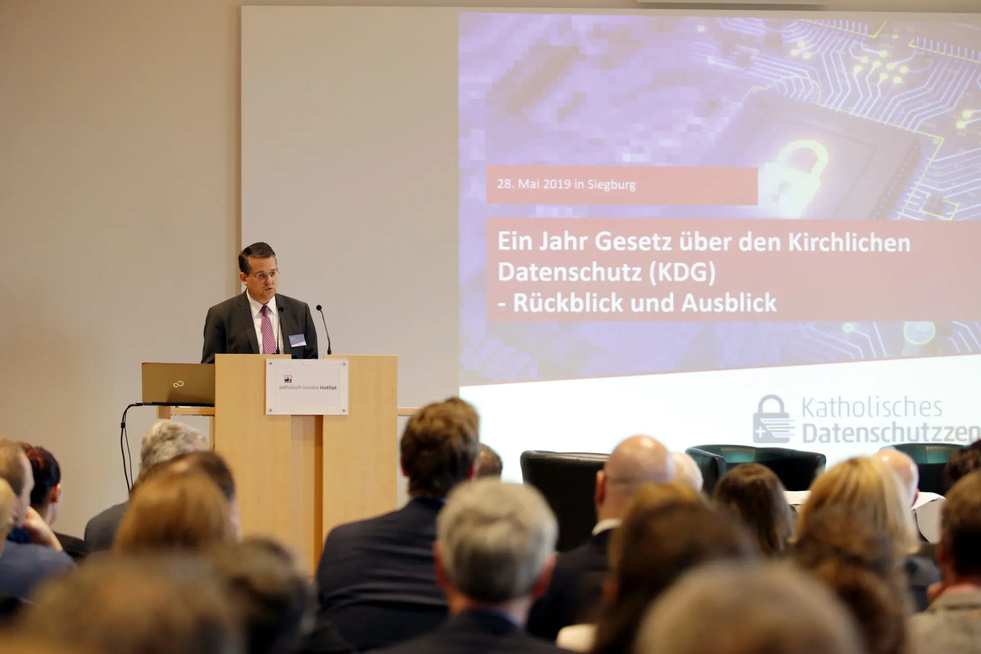 Mann spricht auf einer Konferenz über den Kirchlichen Datenschutz (KDG) mit Präsentationsfolien im Hintergrund.