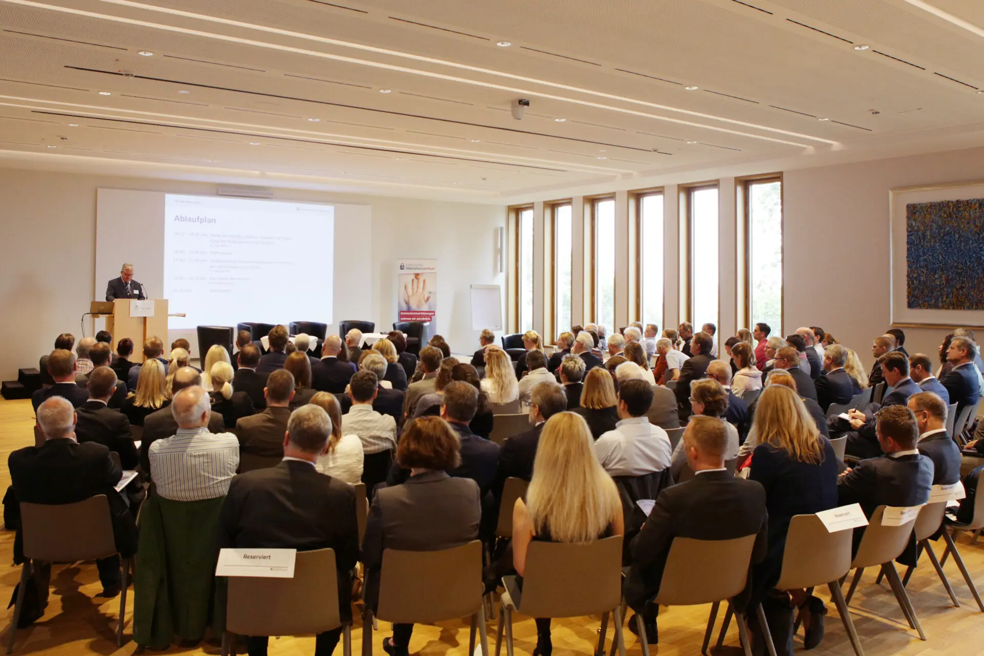 Konferenzraum mit Publikum, das einem Vortragenden auf der Bühne während einer Veranstaltung zuhört.