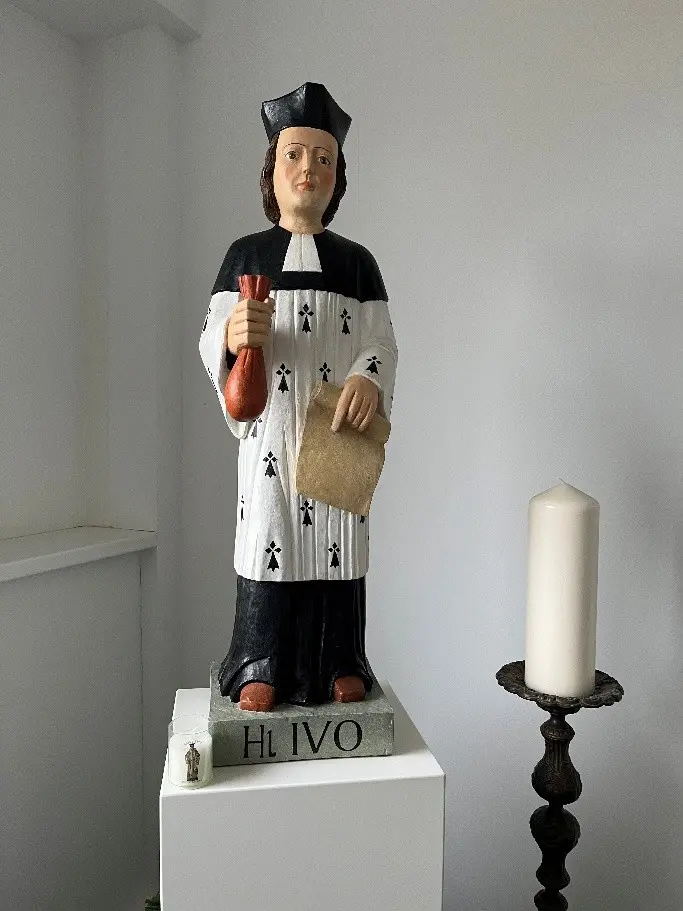 Statue des Heiligen Ivo in traditioneller Kleidung mit einer Urkunde und einem Buch in der Hand, auf einem weißen Sockel platziert.